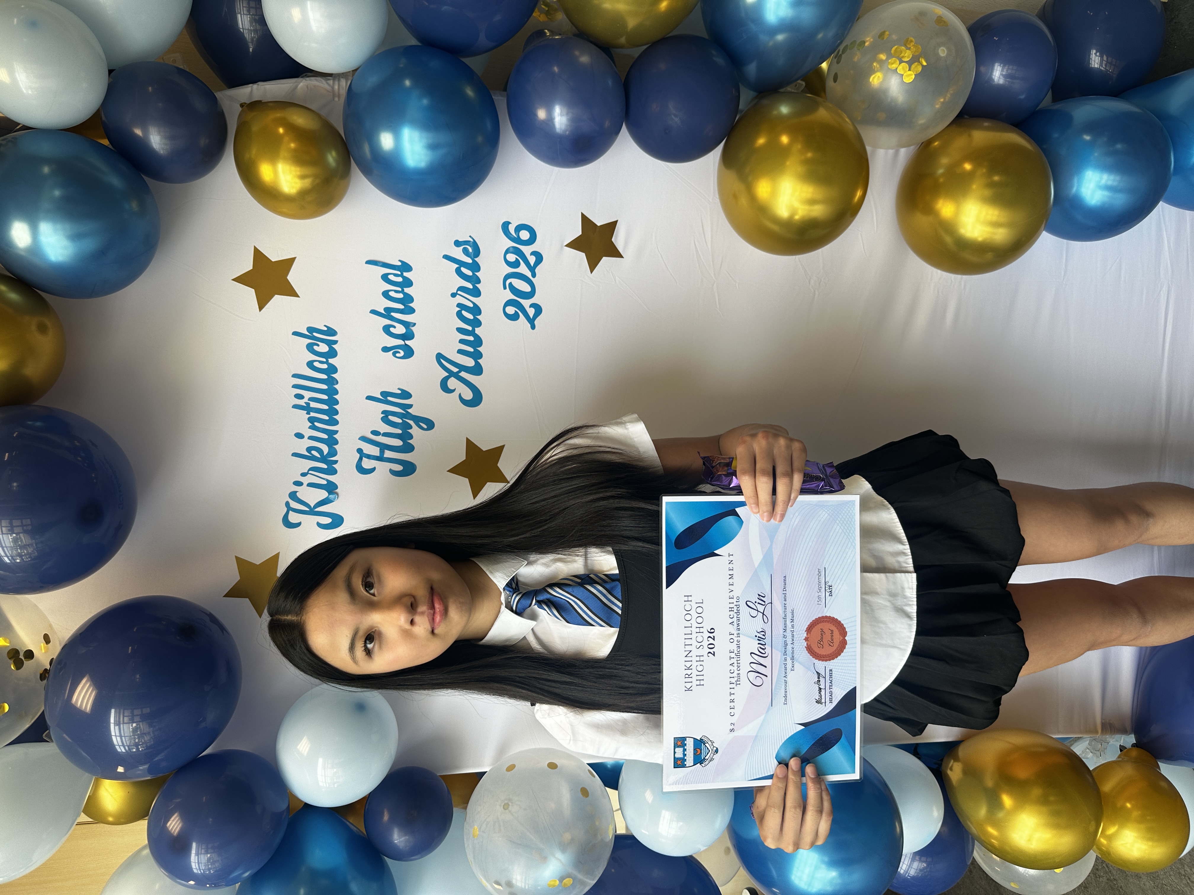 pupil standing in front of a sign and blue baloons while holding a certificate