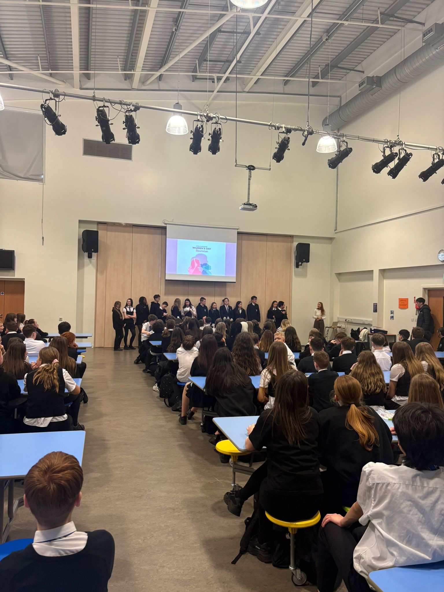 Pupils at assembly, some standing at the front and others sitting at table.