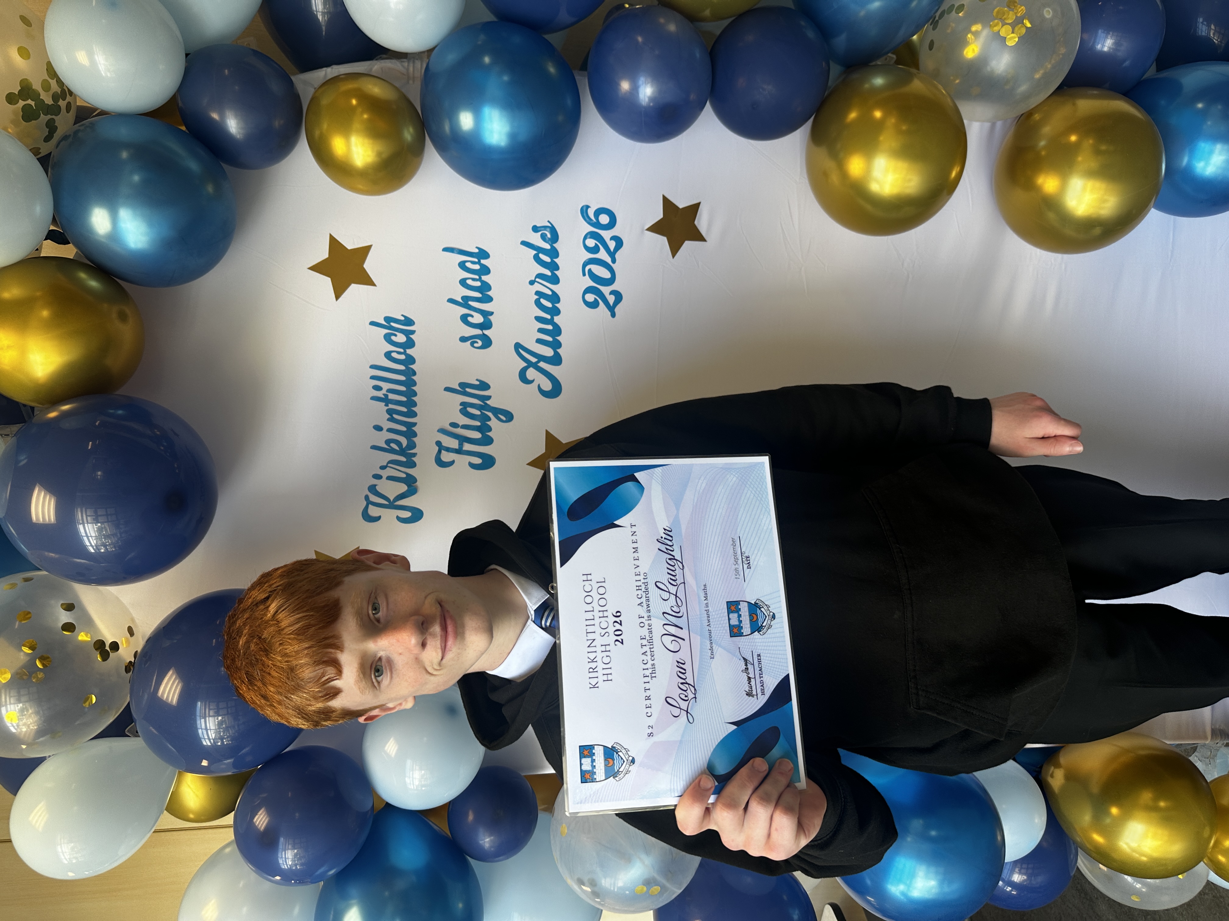 Pupil holding a certificate with balloons and a sign in the background