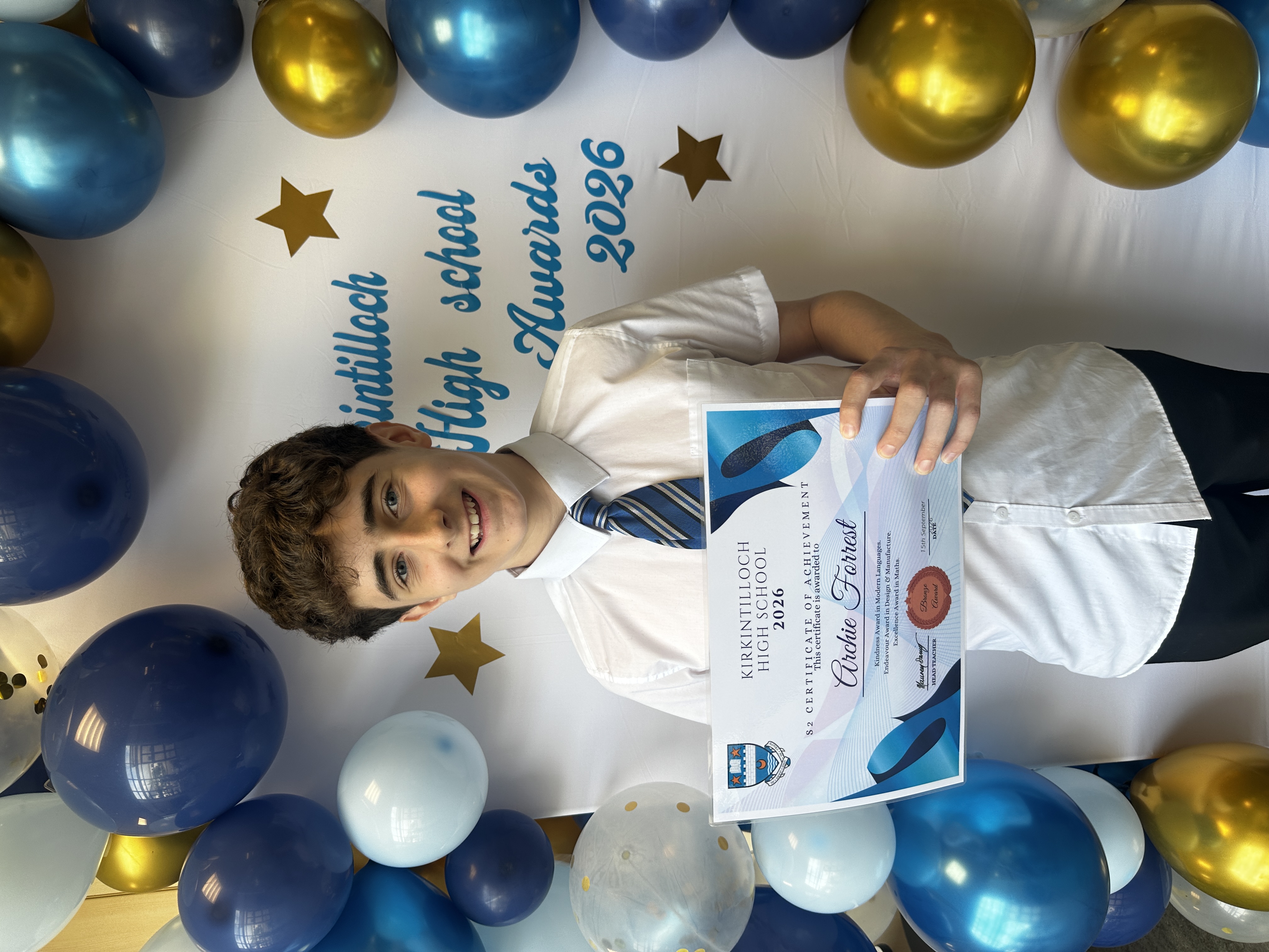 pupil holding a certificate of achievement with blue balloons in the background